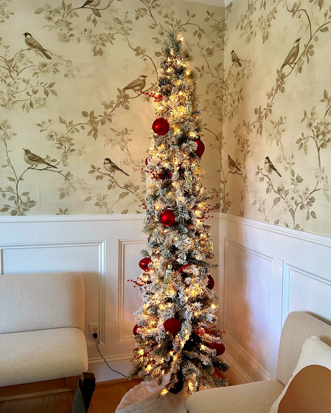 A slim flocked Christmas tree with burgundy ornaments in a wallpapered sitting room